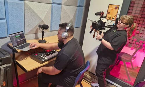 Two people creating digital content, one sitting at the computer, the other standing behind him with a video camera