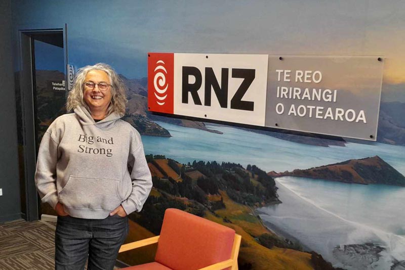 Kim Morton stands alongside RNZ's logo on the wall at RNZ's Christchurch office