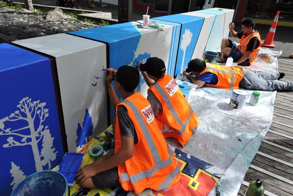 Four artists at Kākano Youth Arts Collective, wearing orange high-vis vests and focused on painting a mural on a concrete wall